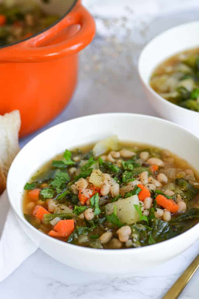 Close-up of a bowl of Italian White Bean and escarole soup with an orange dutch oven in the background
