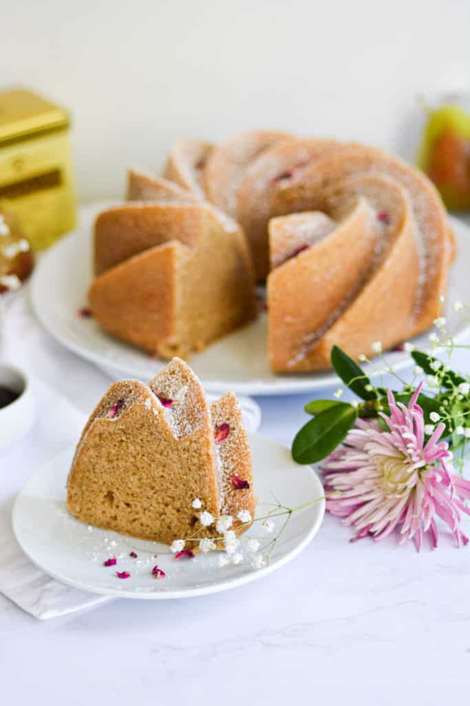 A slice of Bundt cake on a white plate next to puruple flowers