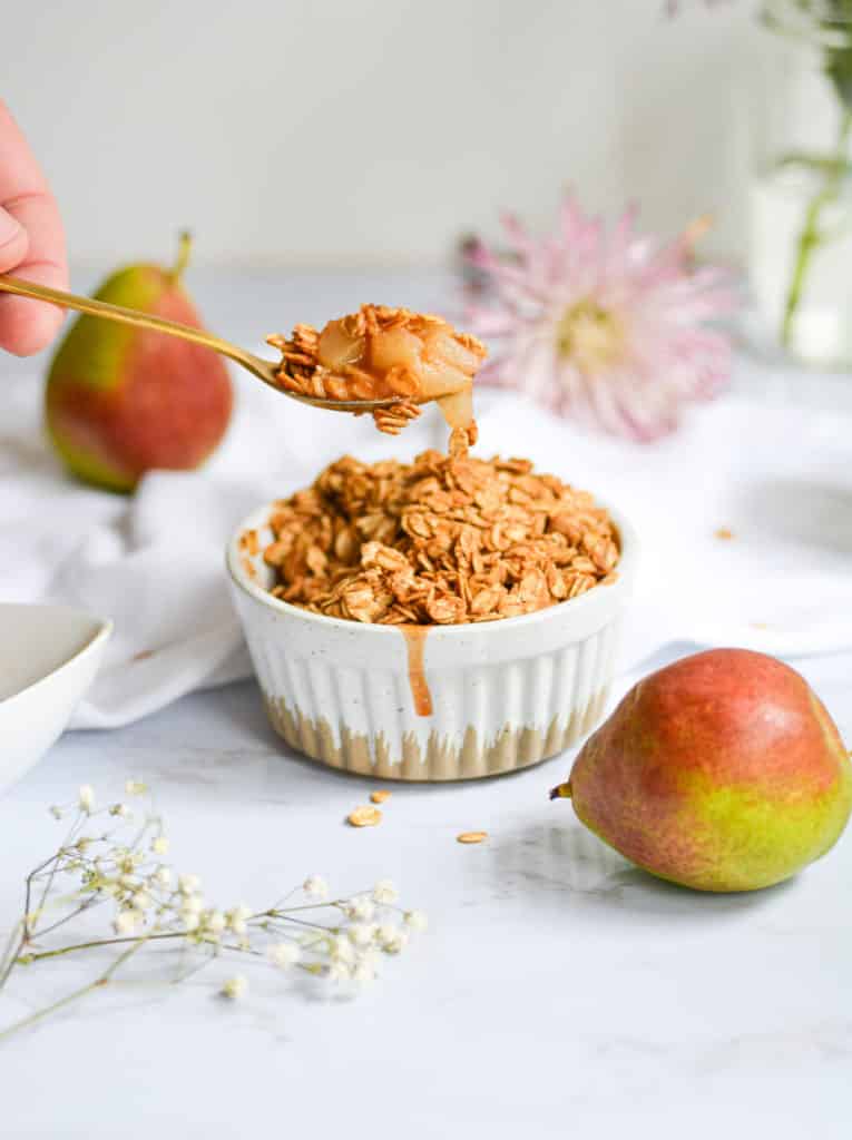 Spoon with a bite of vegan pear crumble on it with a white ramekin and small pears in the background