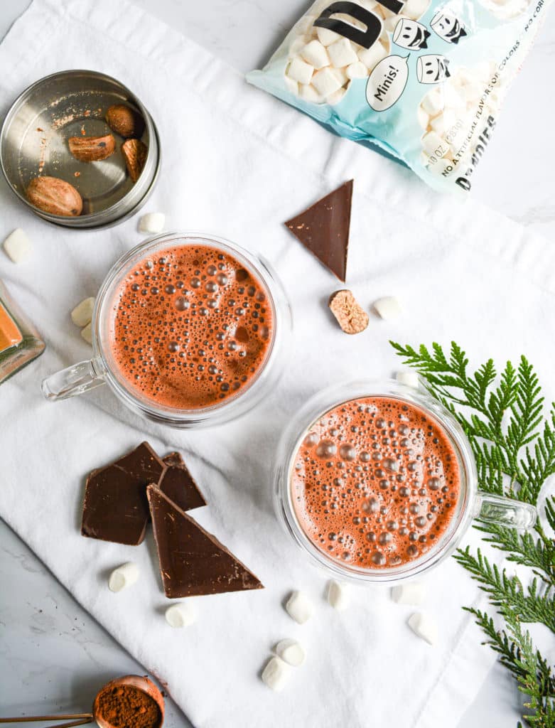 over head shot of two mugs of hot cocoa with nutmeg and greenery in the background