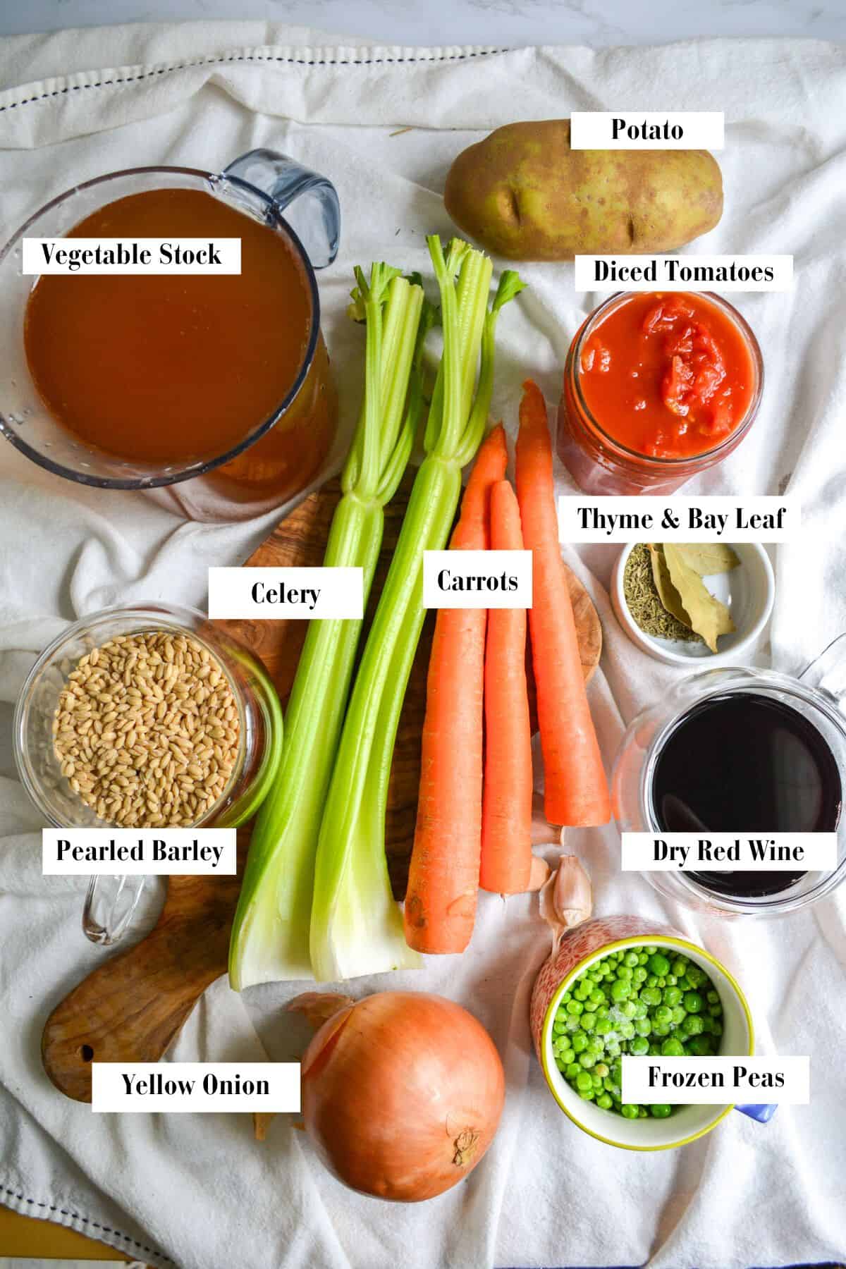 Overhead photo of carrots, celery, an onion, a cup of barley, wine tomatoes peas and herbs.