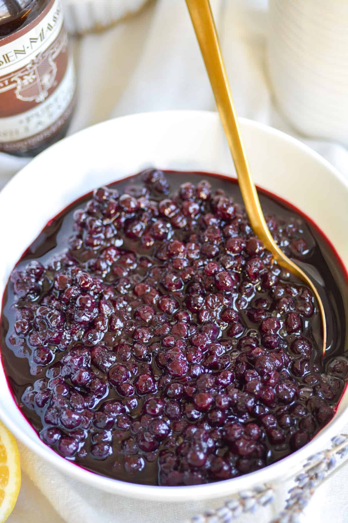 Blueberry Compote recipe in a white bowl with a gold spoon in it.