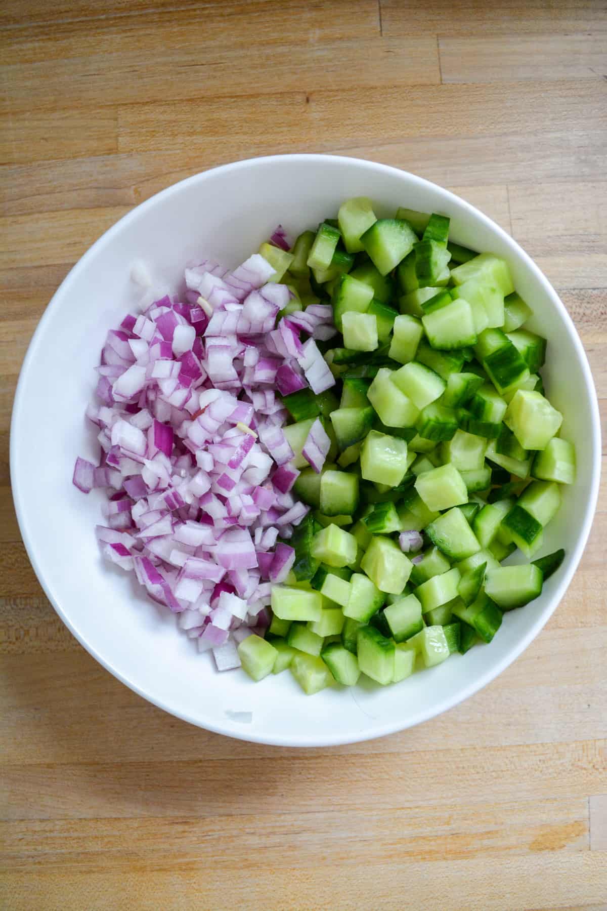 Diced cucumber and red onion in a white bowl on a wooden surface.