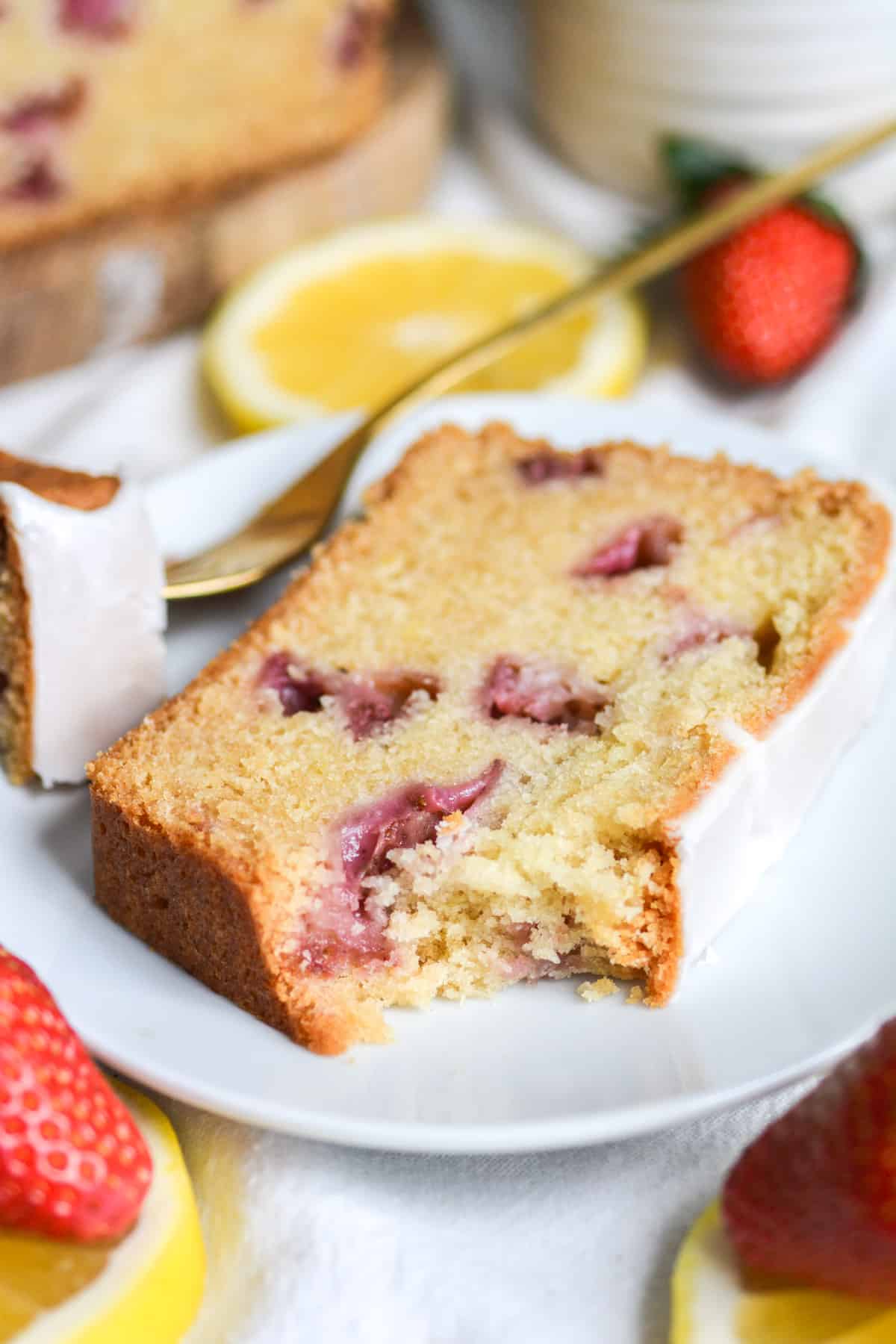 A bite taken out of a slice of vegan strawberry pound cake on a white plate with lemon slices and strawberries in the foreground.