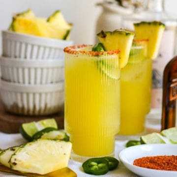 Two glasses of Pineapple Jalapeño Margarita on a linen cloth with slices on pineapple and jalapeño on the table.