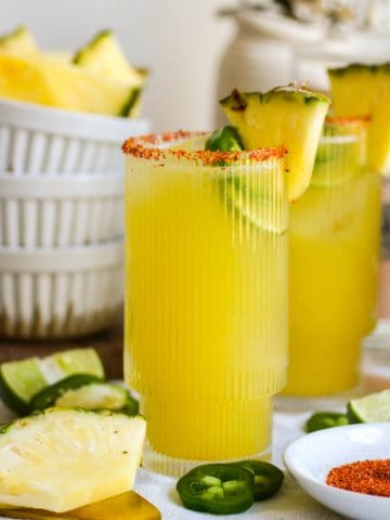 Two glasses of Pineapple Jalapeño Margarita on a linen cloth with slices on pineapple and jalapeño on the table.