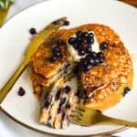 Vegan Blueberry Oat Flour Pancakes on a beige plate with a gold fork and knife with a bite of pancake on the work.
