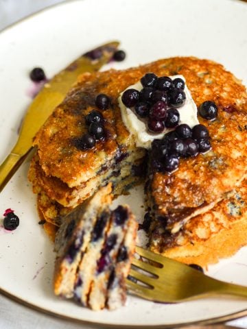 Vegan Blueberry Oat Flour Pancakes on a beige plate with a gold fork and knife with a bite of pancake on the work.