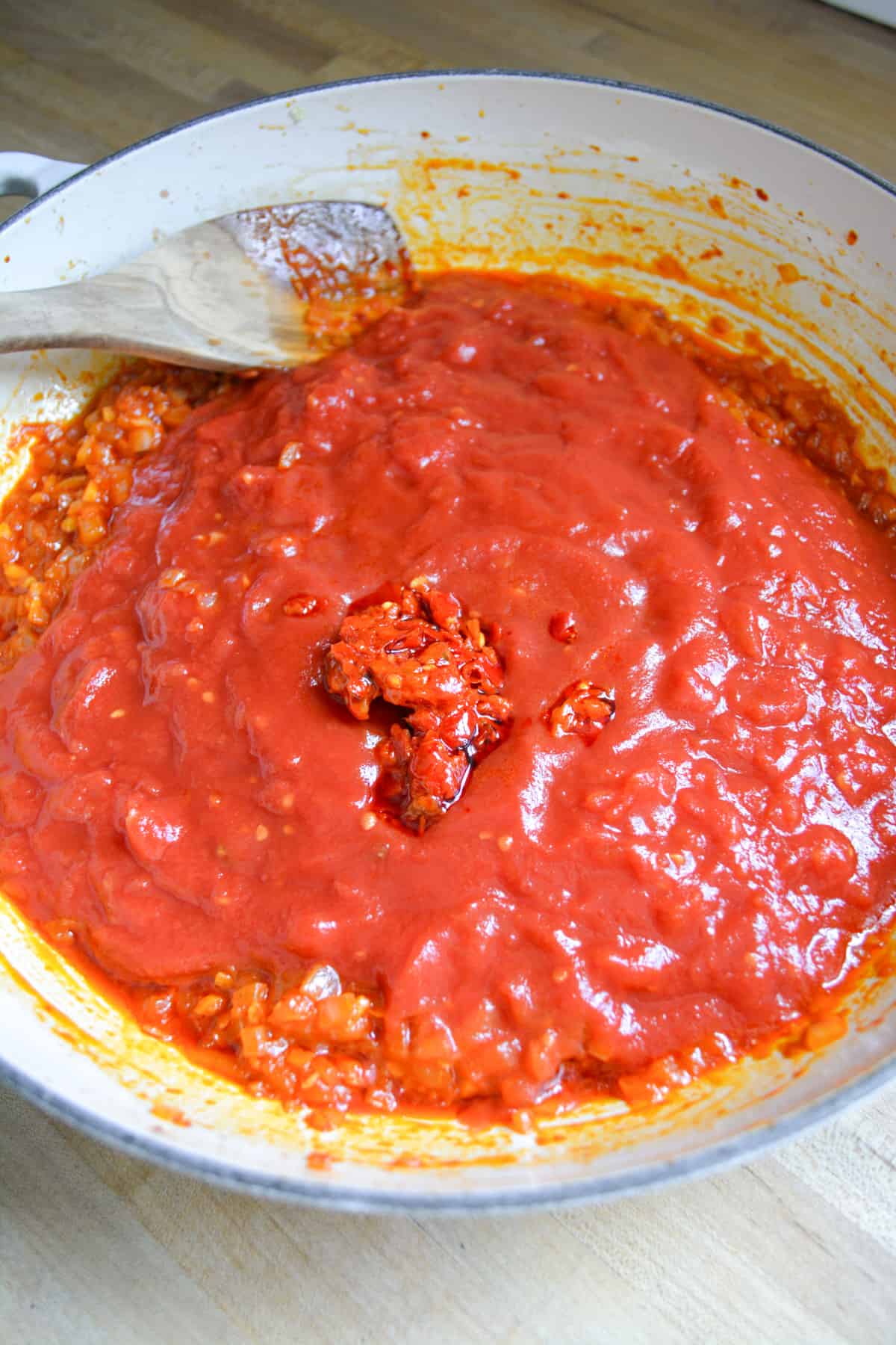 Crushed tomatoes and crushed Calabrian chilis added in to a large skillet with a wooden spoon on the top left.
