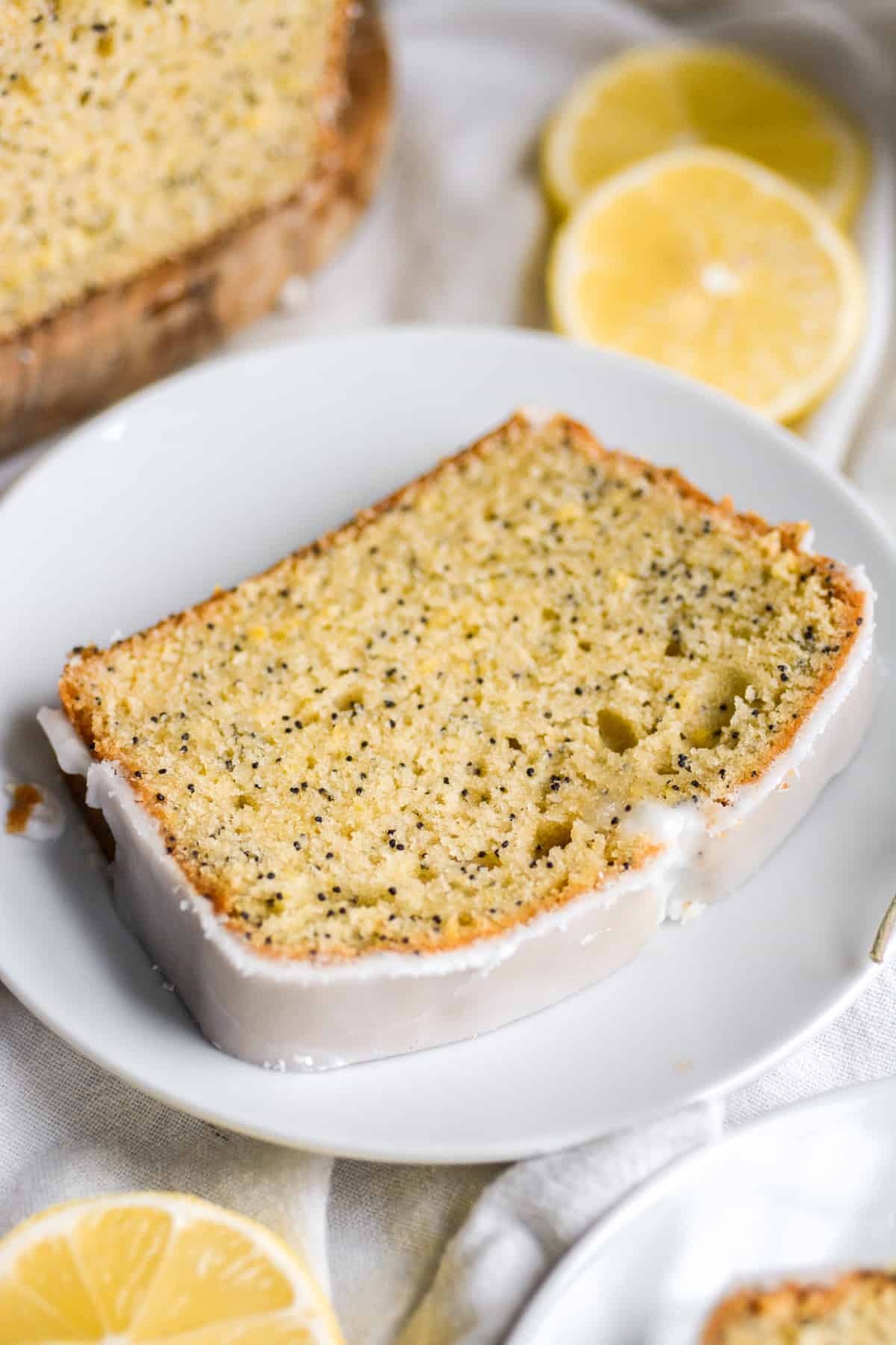A slice of Vegan Lemon Poppyseed Pound Cake  on a white plate with lemon slices in the background.