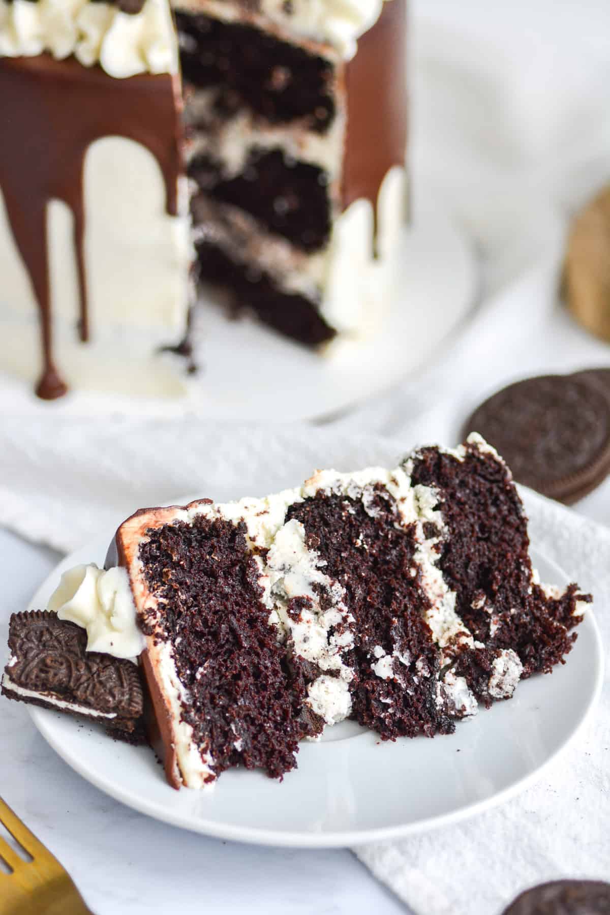 A slice of vegan Oreo drip cake on a white plate with the rest of the cake in the background.