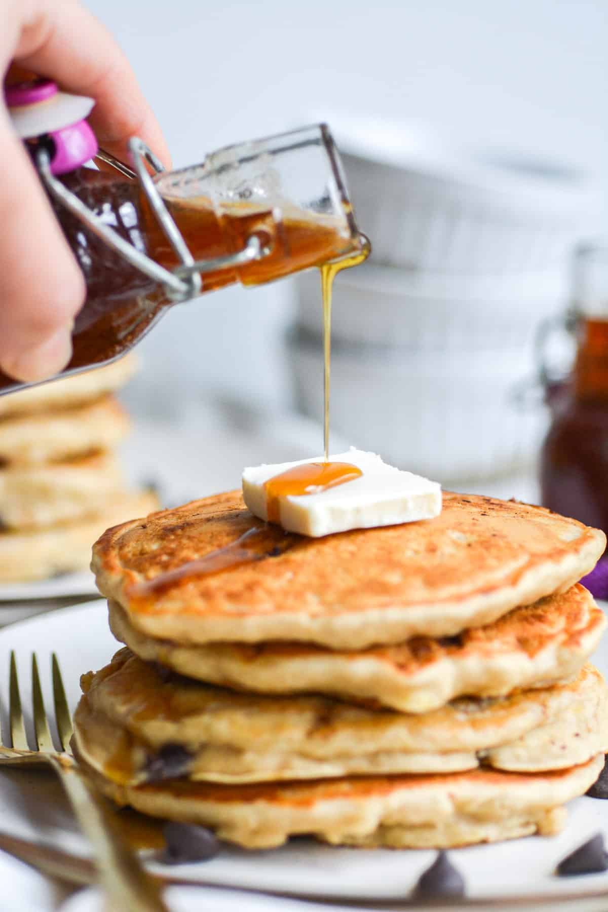 hand pouring maple syrup onto a stack of vegan banana chocolate chip pancakes