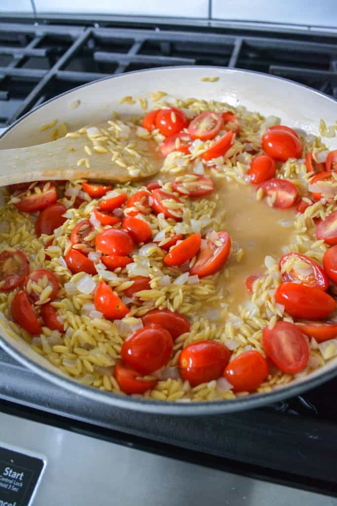 halved cherry tomatoes added into a pan with orzo and some vegetable stock