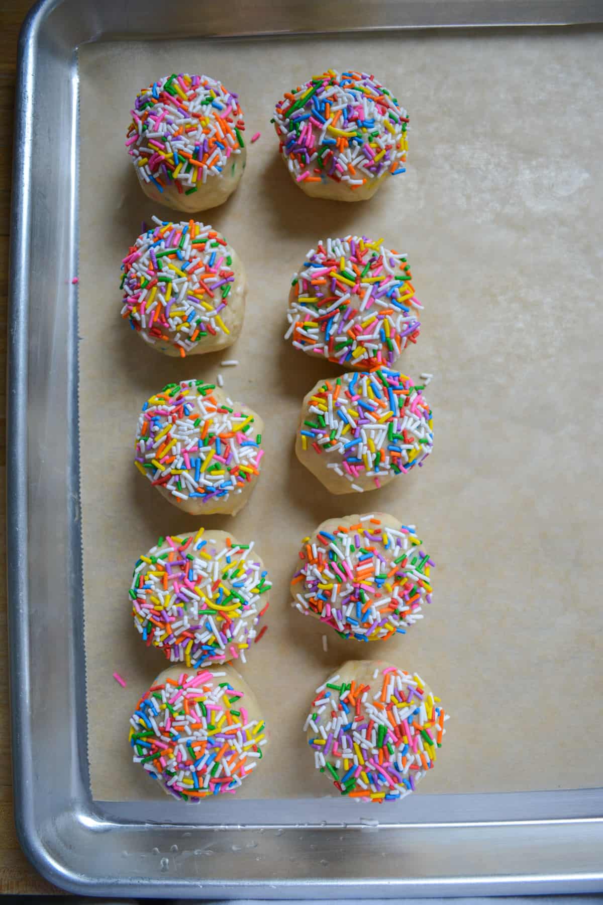 Scooped vegan cookie dough dipped into rainbow sprinkles on a baking sheet.