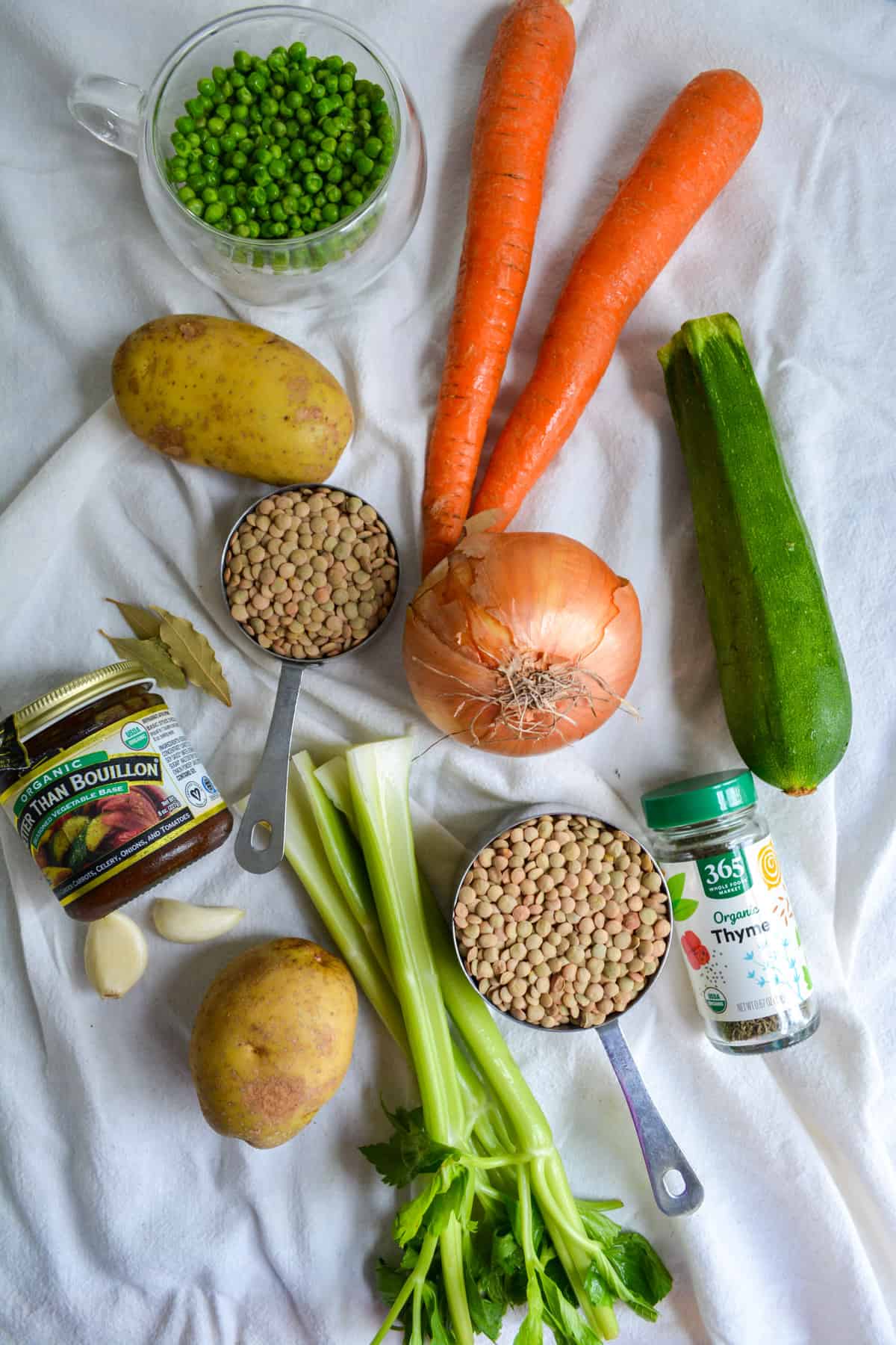 Overhead shot of celery, two carrots, vegetable boullion, lentils, a zucchini, an onion, a cup of peas and a container of thyme.