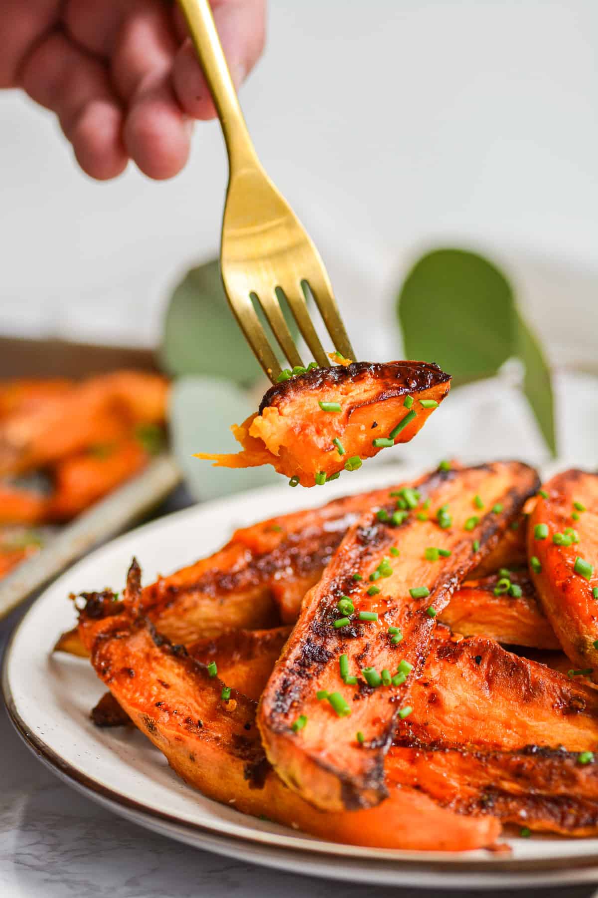A gold fork with a bite of hot honey sweet potatoes on it with a plate of potatoes in the background.
