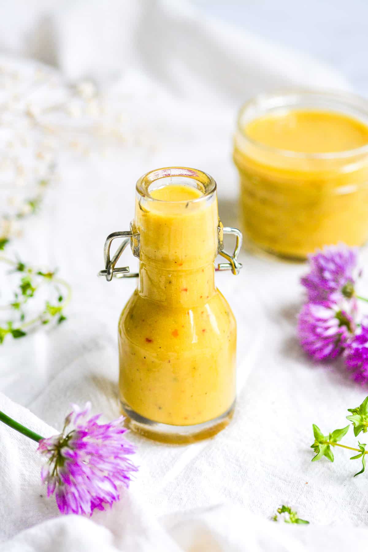 Vegan Honey Mustard Dressing in a small jar on a white cloth with purple flowers in the foreground.