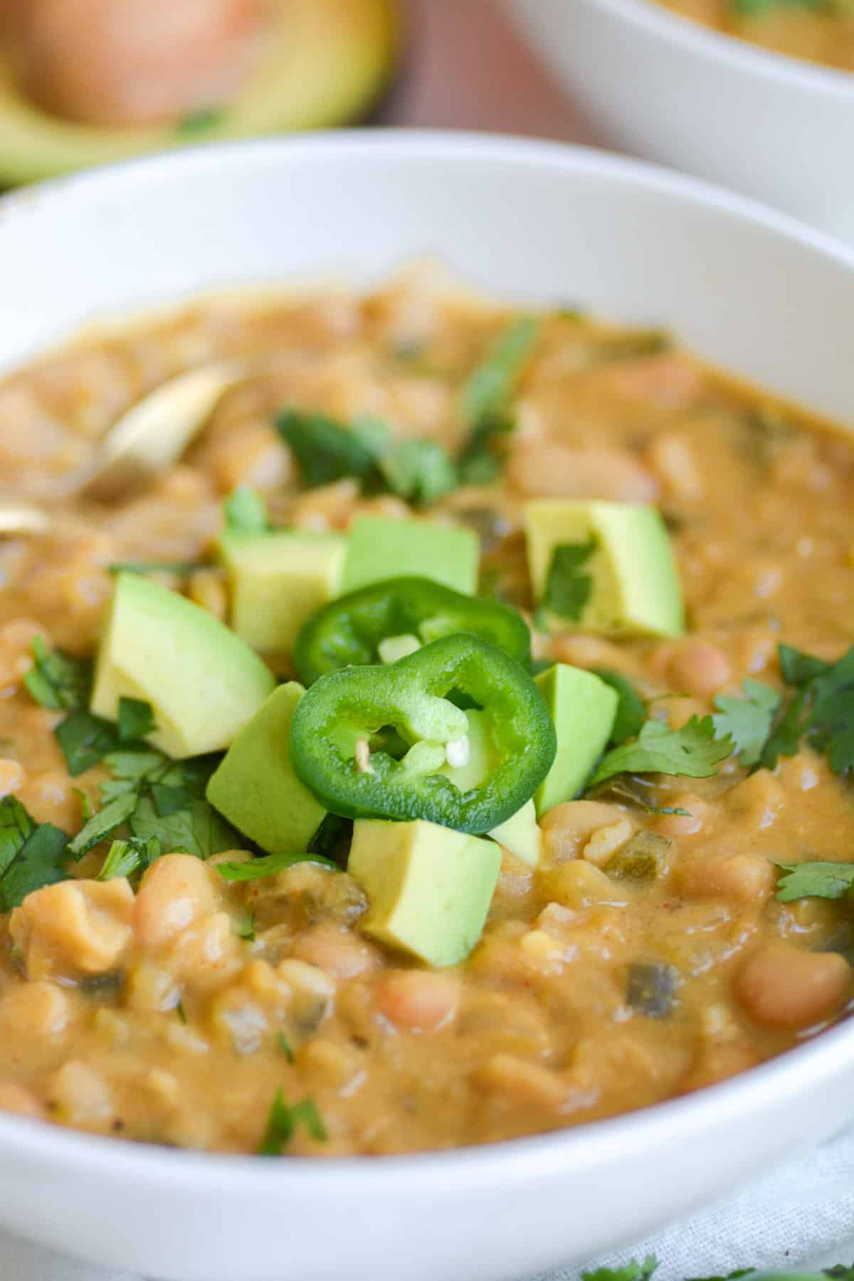 Close up of vegan and vegetarian white bean chili in a bowl topped with avocado and sliced jalapeños.