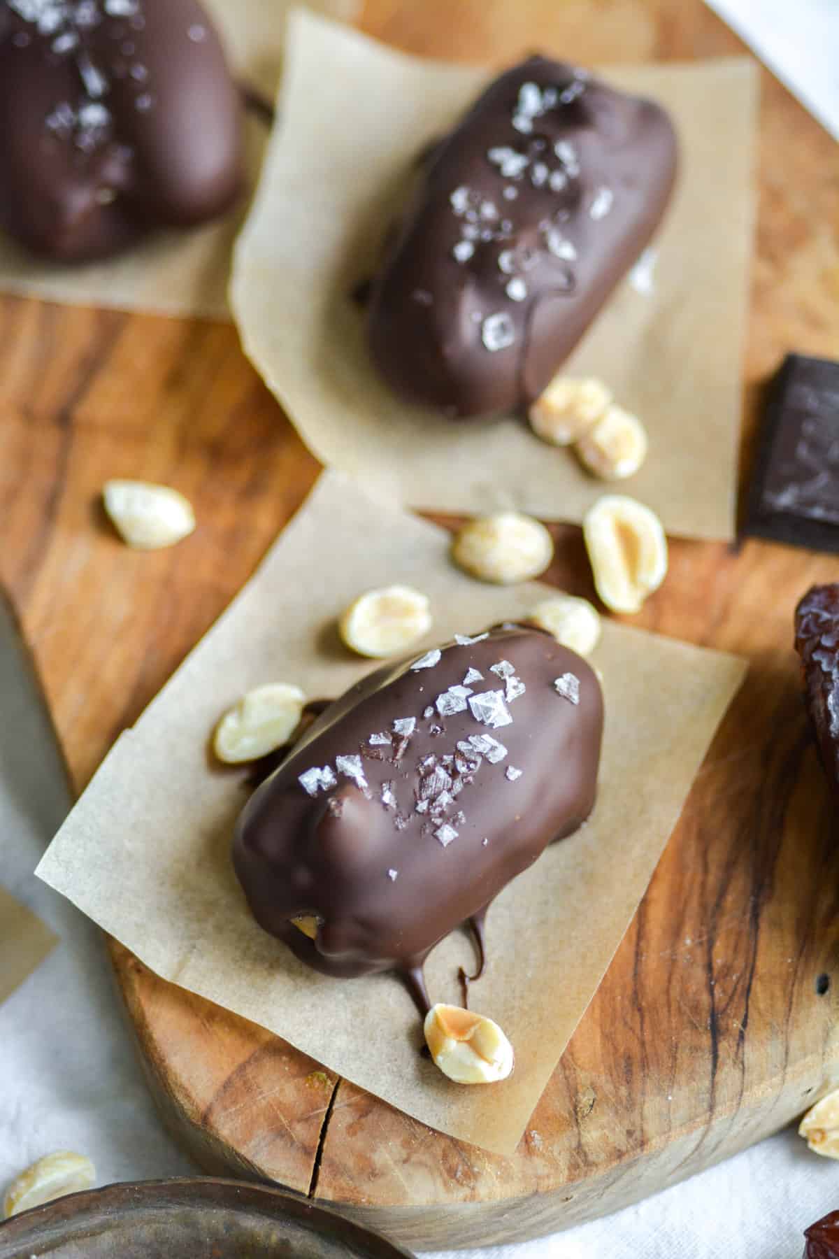 Chocolate covered dates topped with flakey salt on a wooden board with peanuts in the scene.