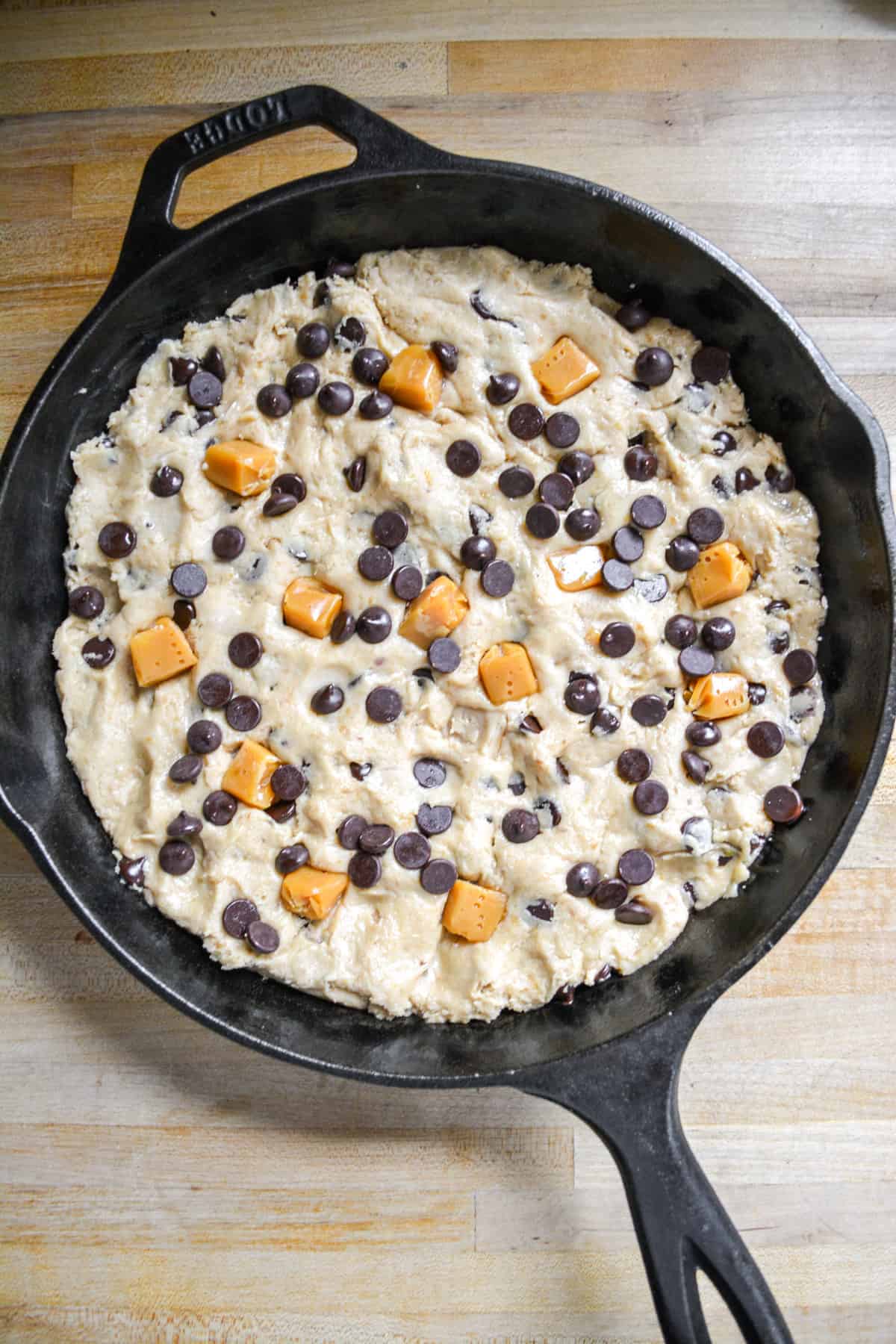 A black cast iron skillet with cookie dough pressed into it topped with chocolate chips and salted caramel candies.