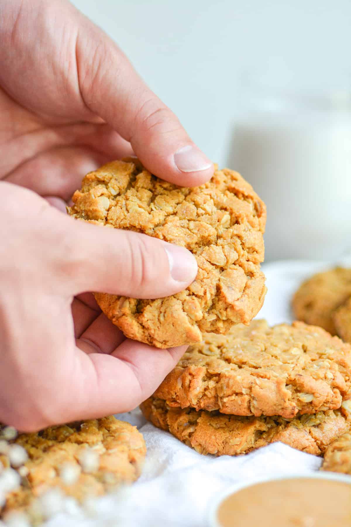 Hands holding a vegan oatmeal peanut butter cookie with more cookies in the foreground.