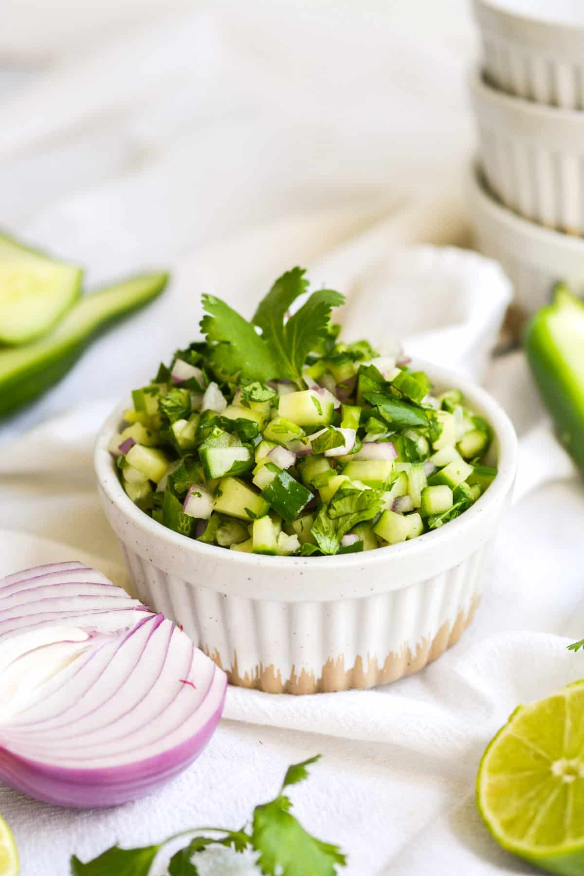 Cucumber pico de gallo in a small bowl with red onion and lime in the foreground.