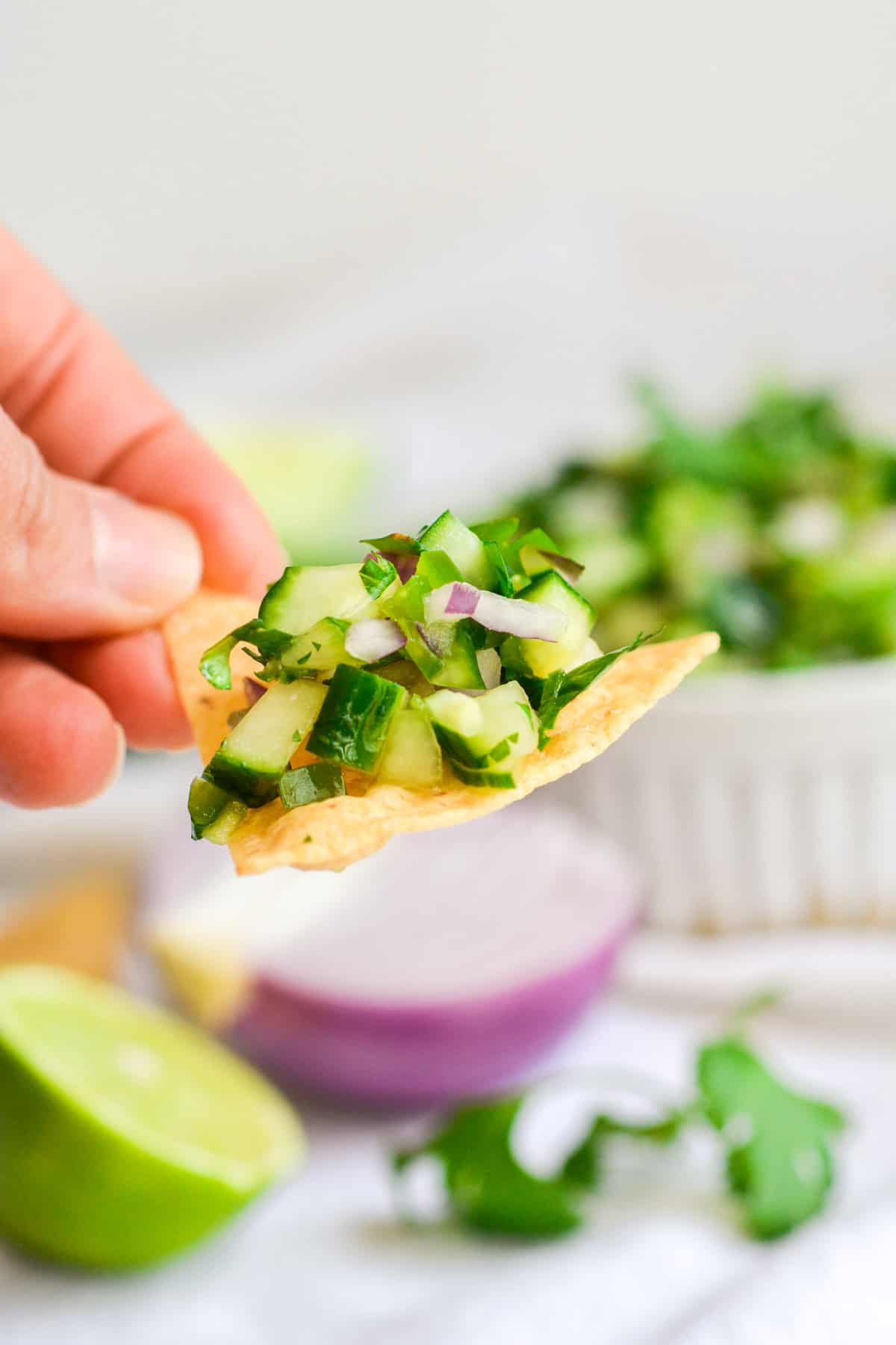 A hand holding a tortilla chip with cucumber pico de gallo on it.