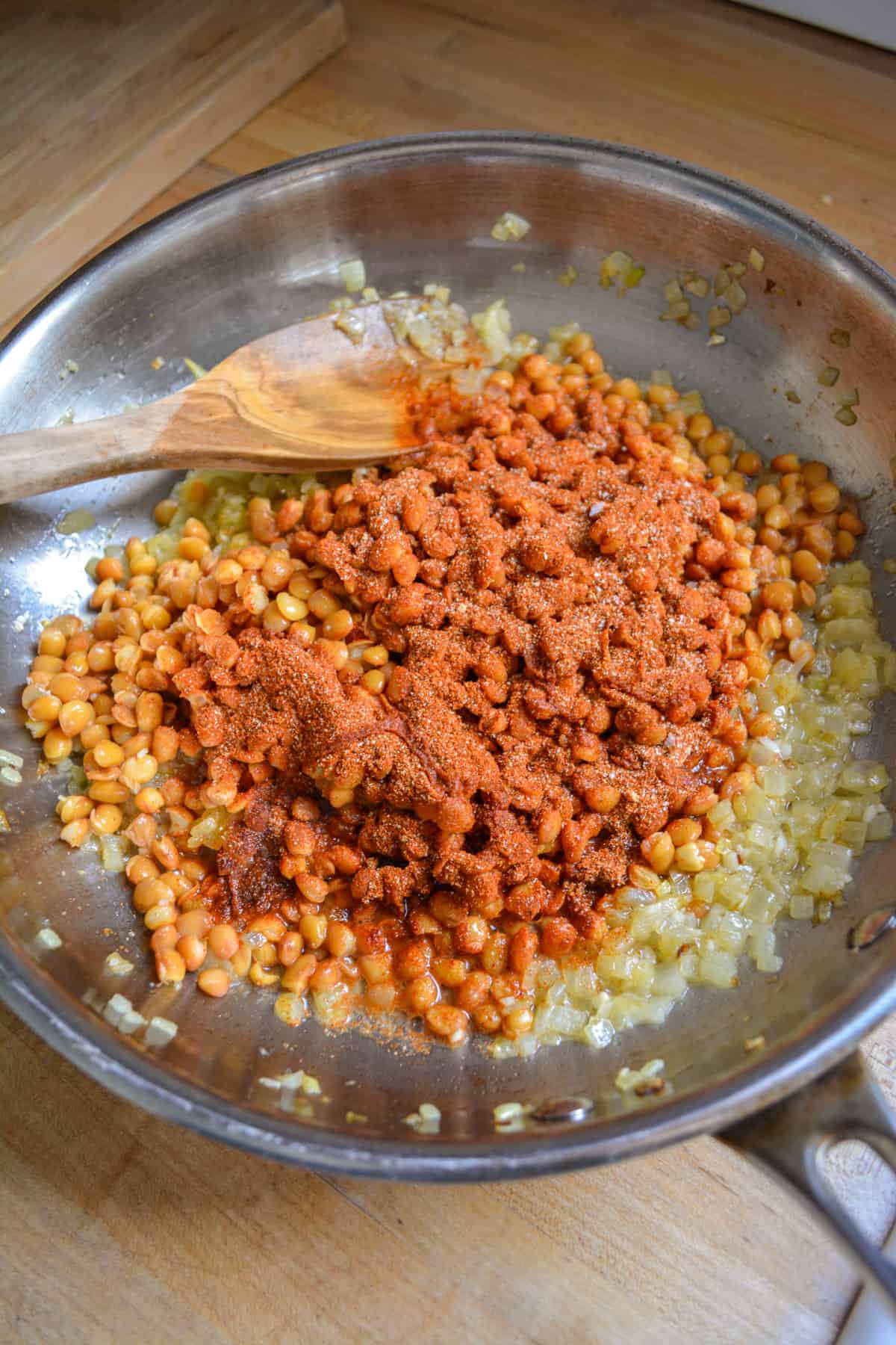 Lentils, spices and water added to a stainless steel skillet with a wooden spoon to the left.