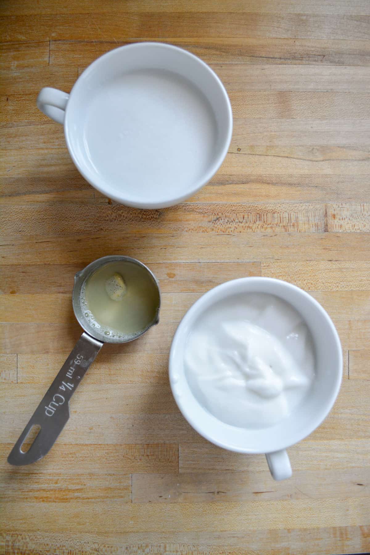 A small cup of vegan yogurt, a small cup of dairy free milk and a small measuring cup of lemon juice on a wooden surface.