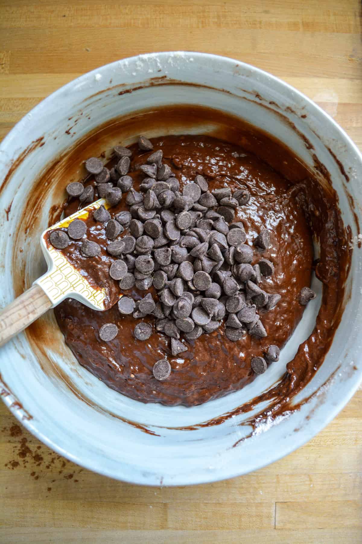 Chocolate chips added into the vegan banana brownie batter in the mixing bowl with a spatula off to the left.