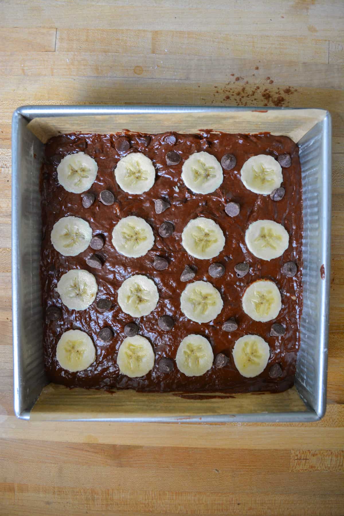 Brownie batter in a parchment-lined baking pan, topped with banana slices and chocolate chips.