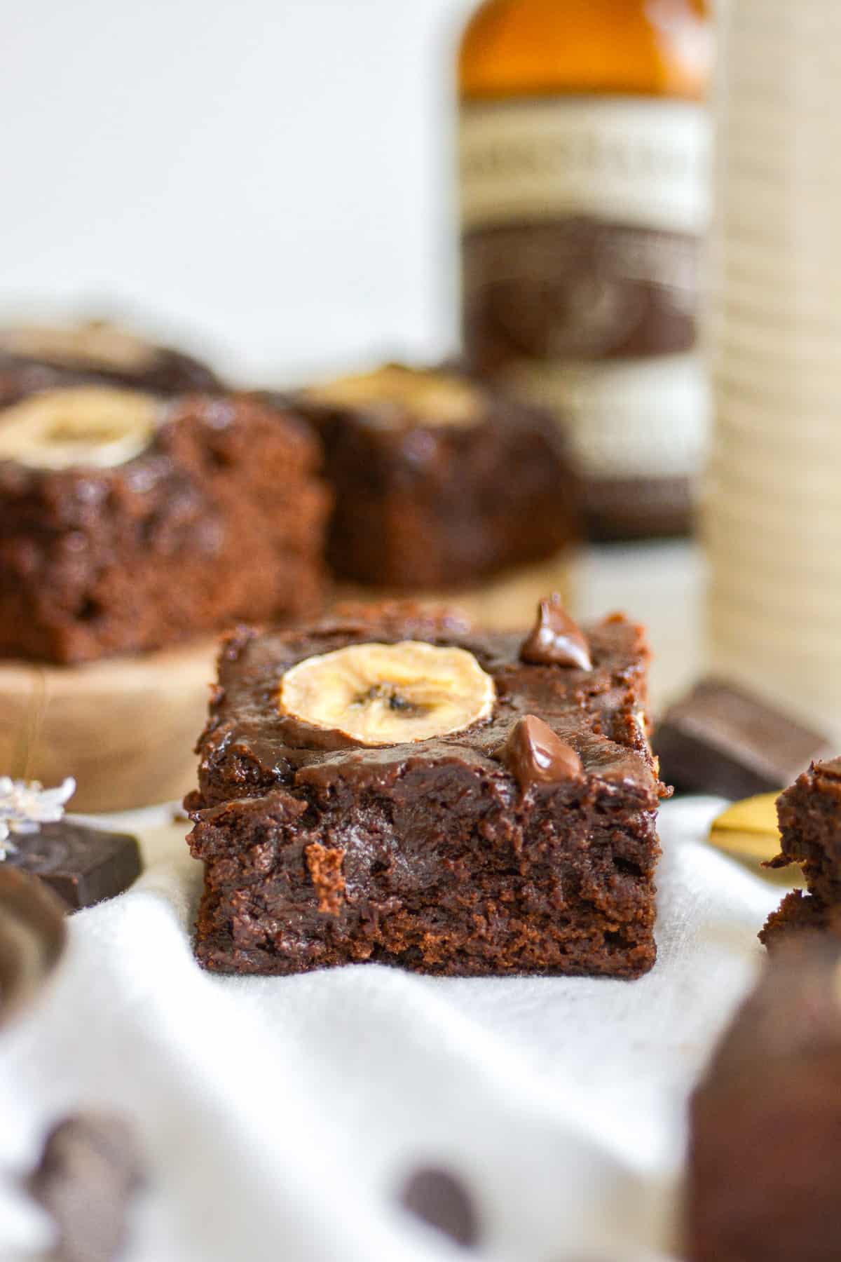 A vegan banana brownie on a white cloth with more brownies on a wooden board in the background.