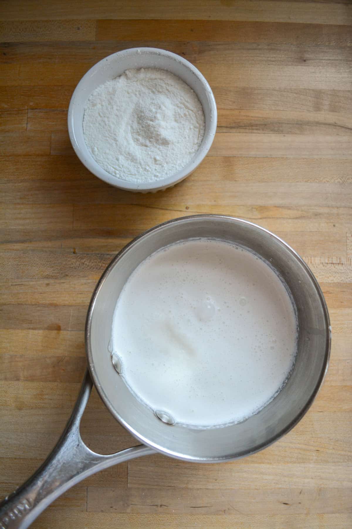 A saucepan with oatmilk and vegan cream in it and a small bowl with cornstarch and sugar mixed together.