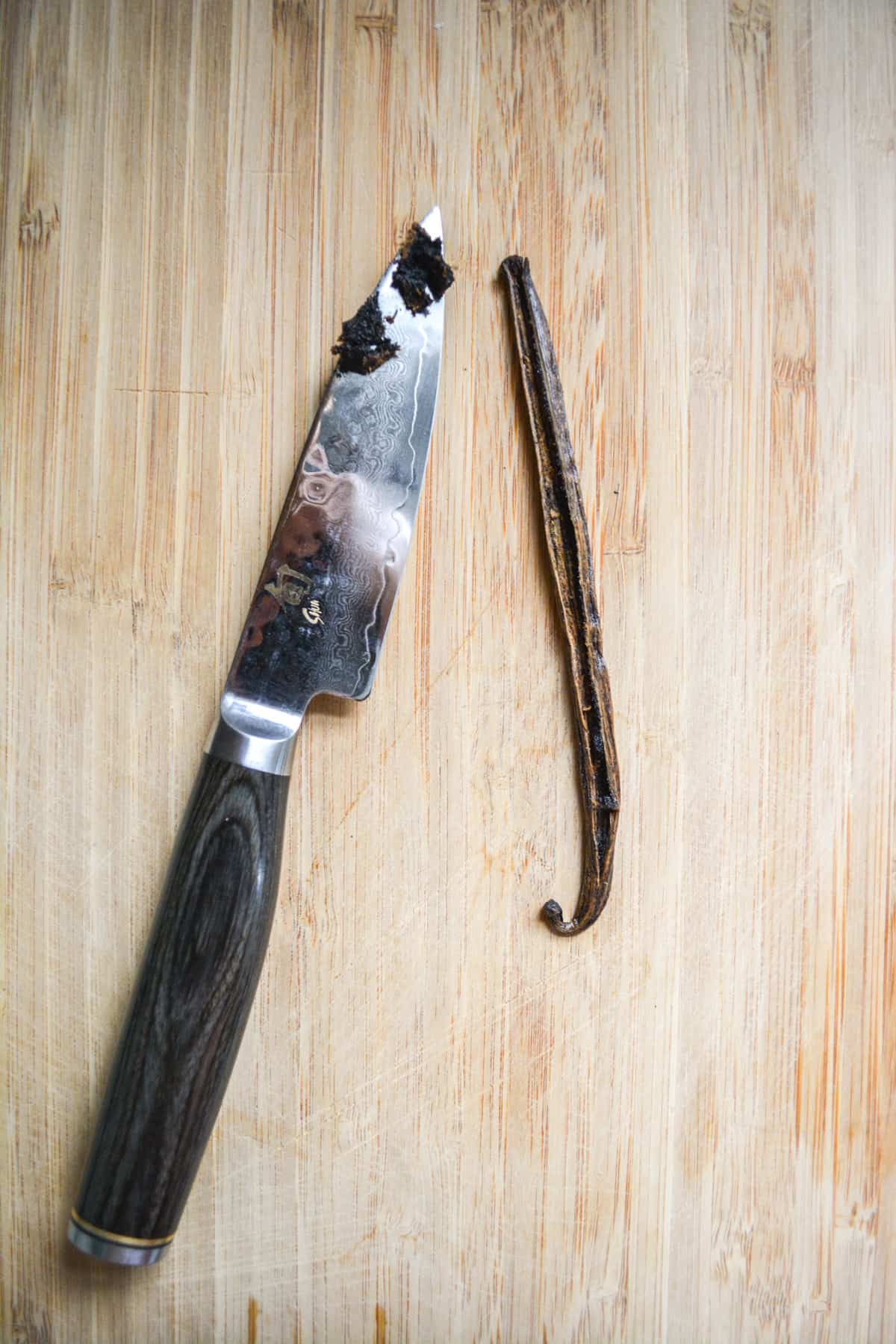 Sliced open vanilla bean on a cutting board next to a paring knife with the vanilla caviar on it.
