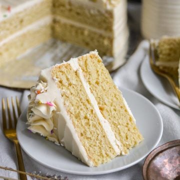 A slice of vegan vanilla layer cake on a plate with a fork to the left and the rest of the cake in the background.