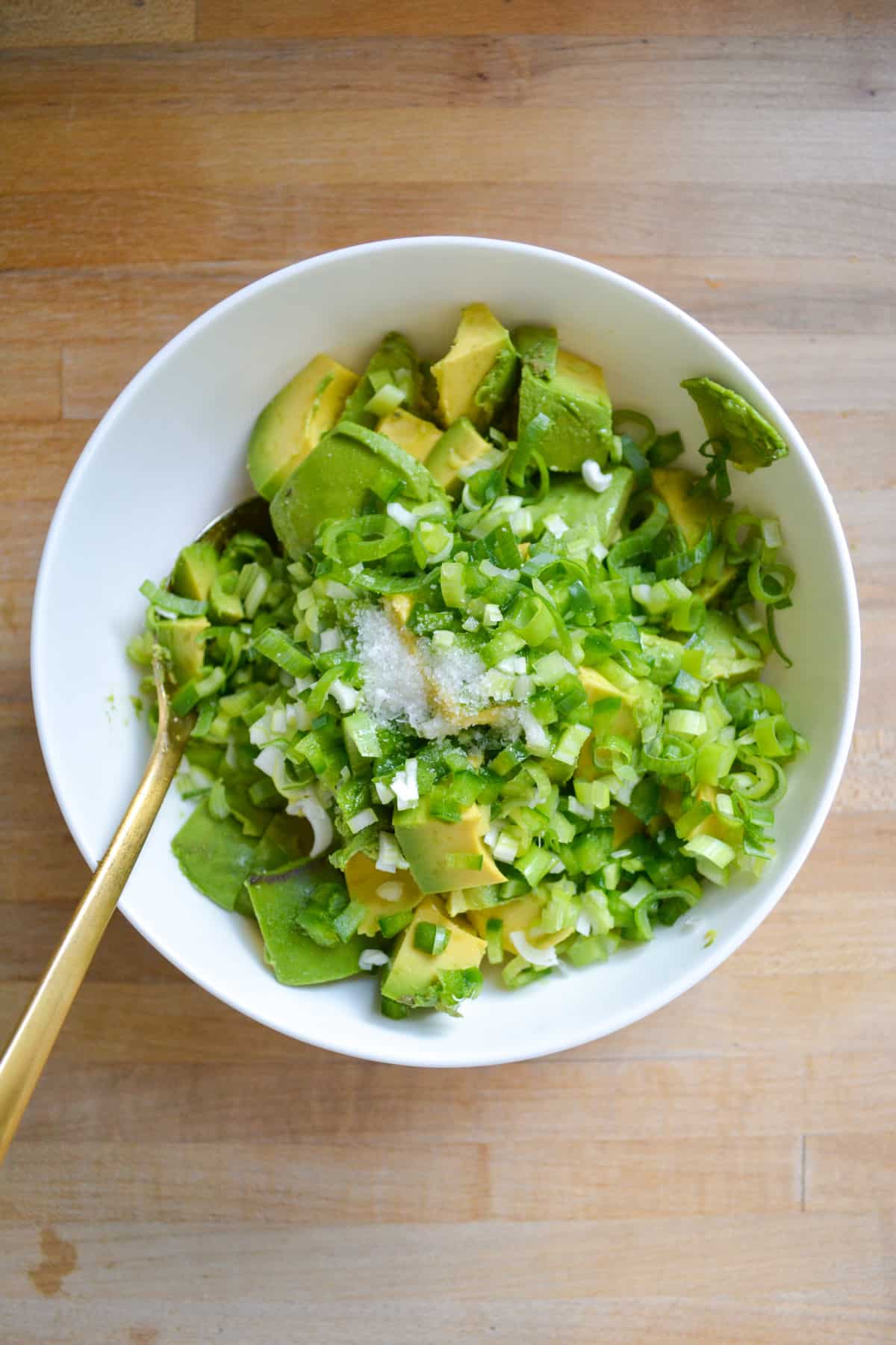 Avocado, jalapeno, spring onion, lime juice and salt in a bowl with a spoon to the left.