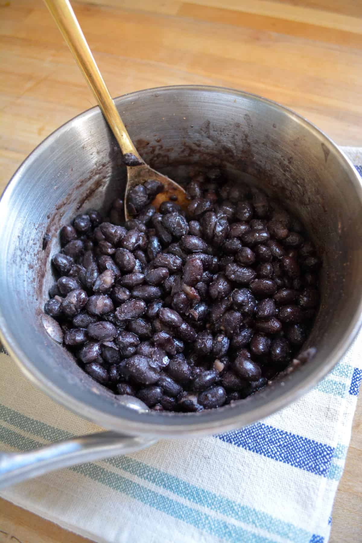 Simmered black beans in a small saucepan with a spoon to the top left.