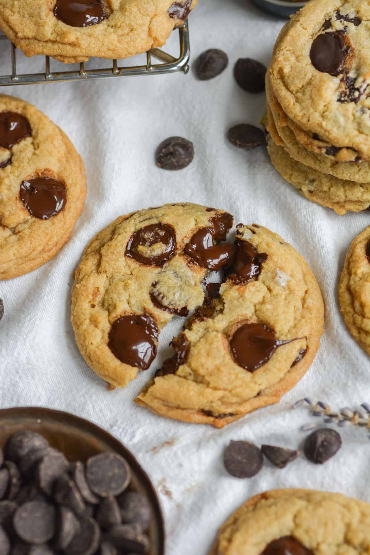 A broken in half Vegan Chocolate Chip Cookie on a white cloth with other cookies and chocolate chips around it. 