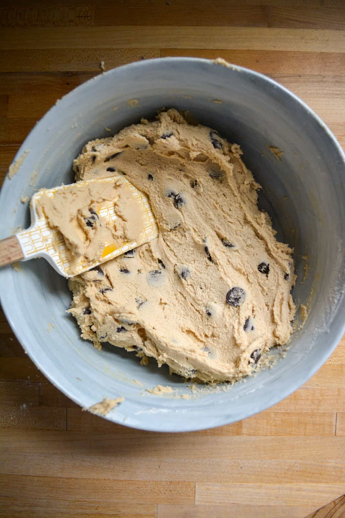 Chocolate chips are mixed into the cookie dough in a mixing bowl with a rubber spatula to the left side of the bowl.