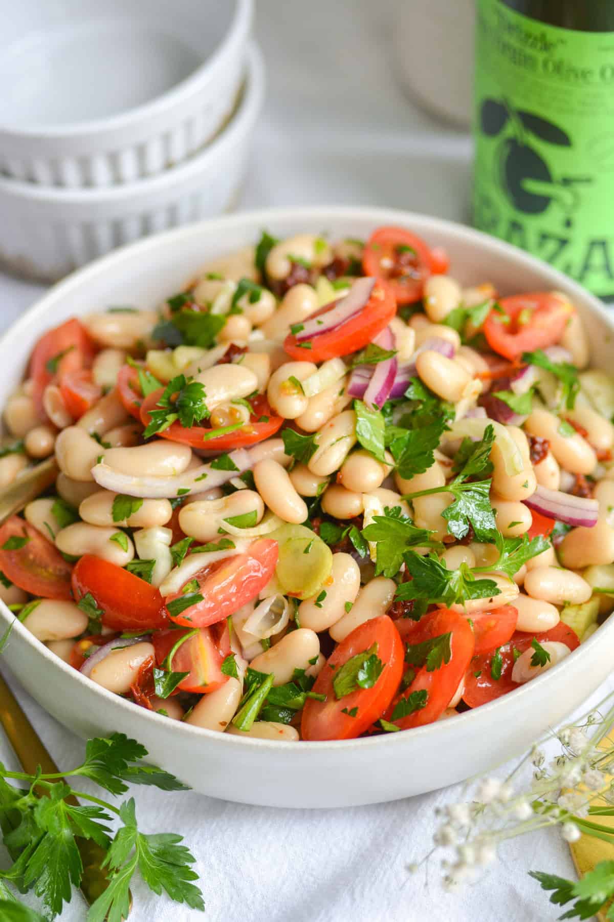 Italian Cannellini Bean Salad in a large bowl on a white cloth with an olive oil bottle in the background.