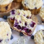 Vegan Lemon Blueberry Muffin is broken in half to show the inside on a paper muffin liner on a white cloth.