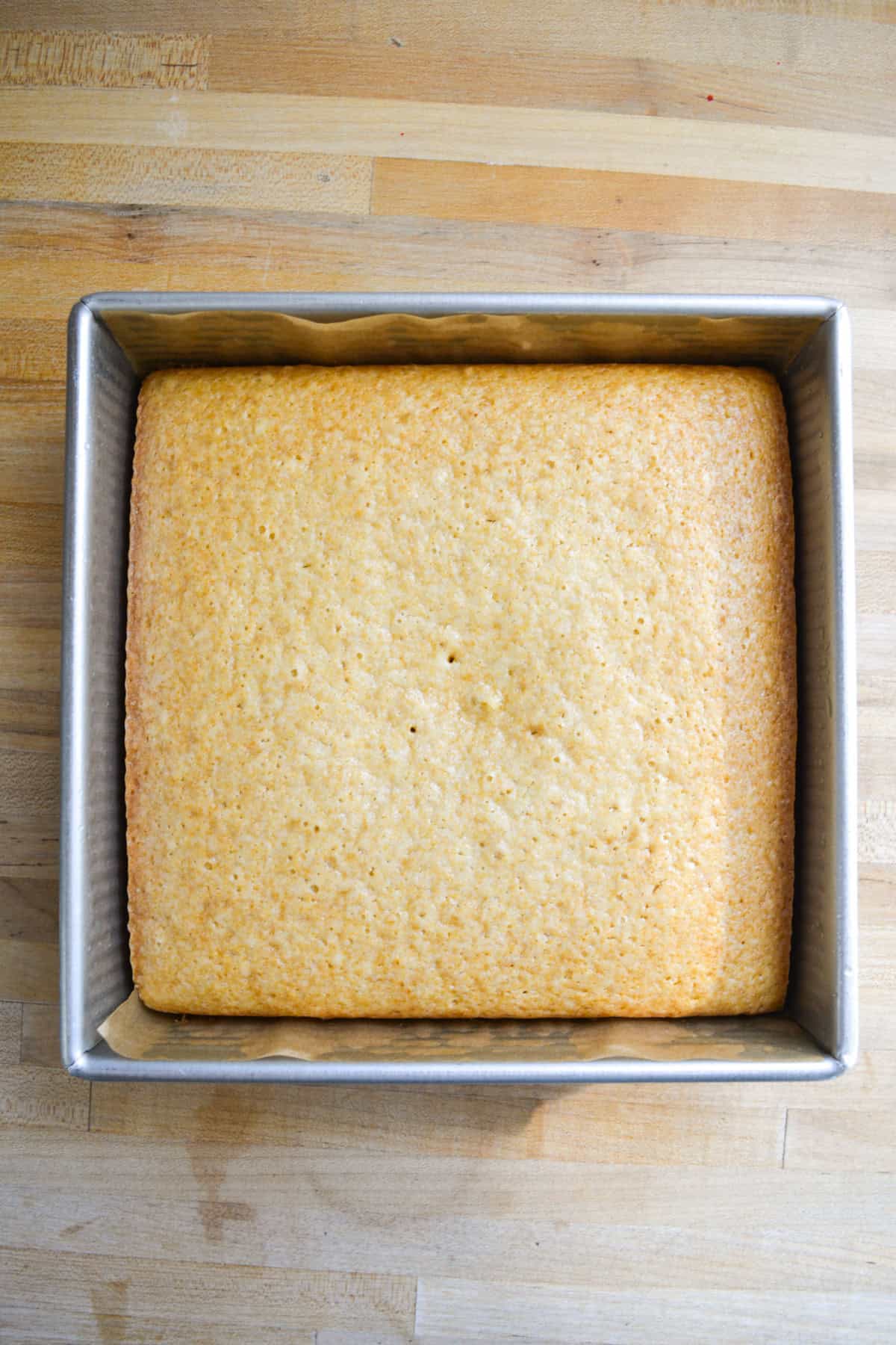 Baked vanilla cake in an 8x8 inch square pan on a wooden surface.