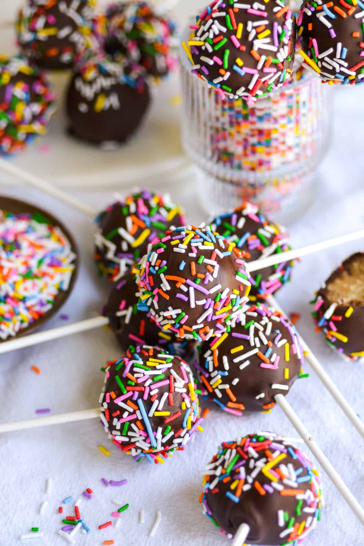 Vegan Cake pops dipped in chocolate and topped with rainbow sprinkles stacked on top of each other on a white cloth.