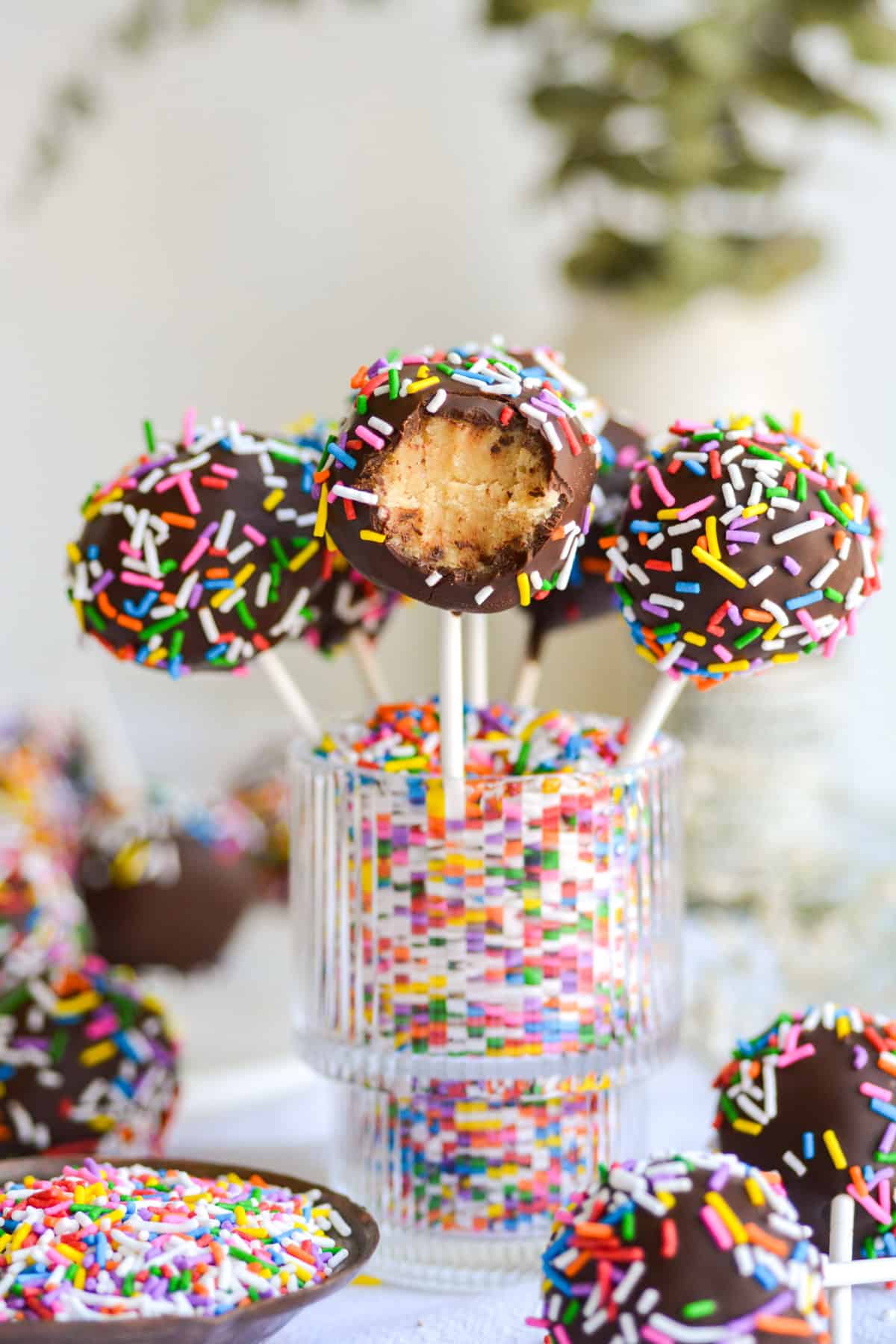 Vegan Cake Pops standing in a glass that is filled with rainbow sprinkles. The cake pop in front has a bite taken out of it.