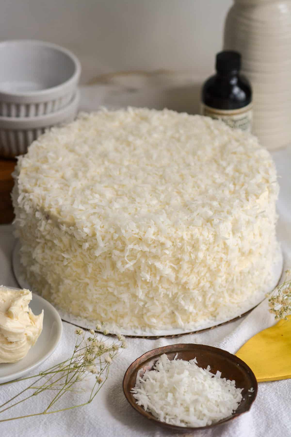 Vegan Coconut Cake covered in coconut on a white cloth with a small bowl of shredded coconut in front of it.