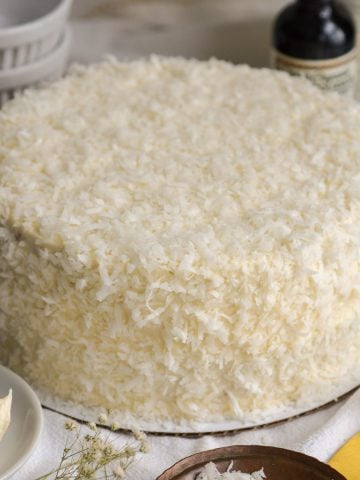 Round Vegan Coconut Cake on a white cloth with a small bowl of coconut in the foreground.