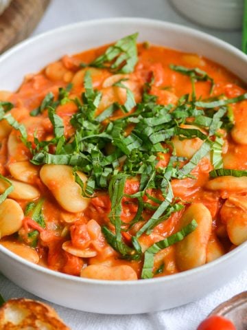 Vegan Creamy Tuscan Butter Beans in a wide shallow bowl topped with sliced basil. The bowl is on a white cloth with a small bowl of tomatoes in the foreground.