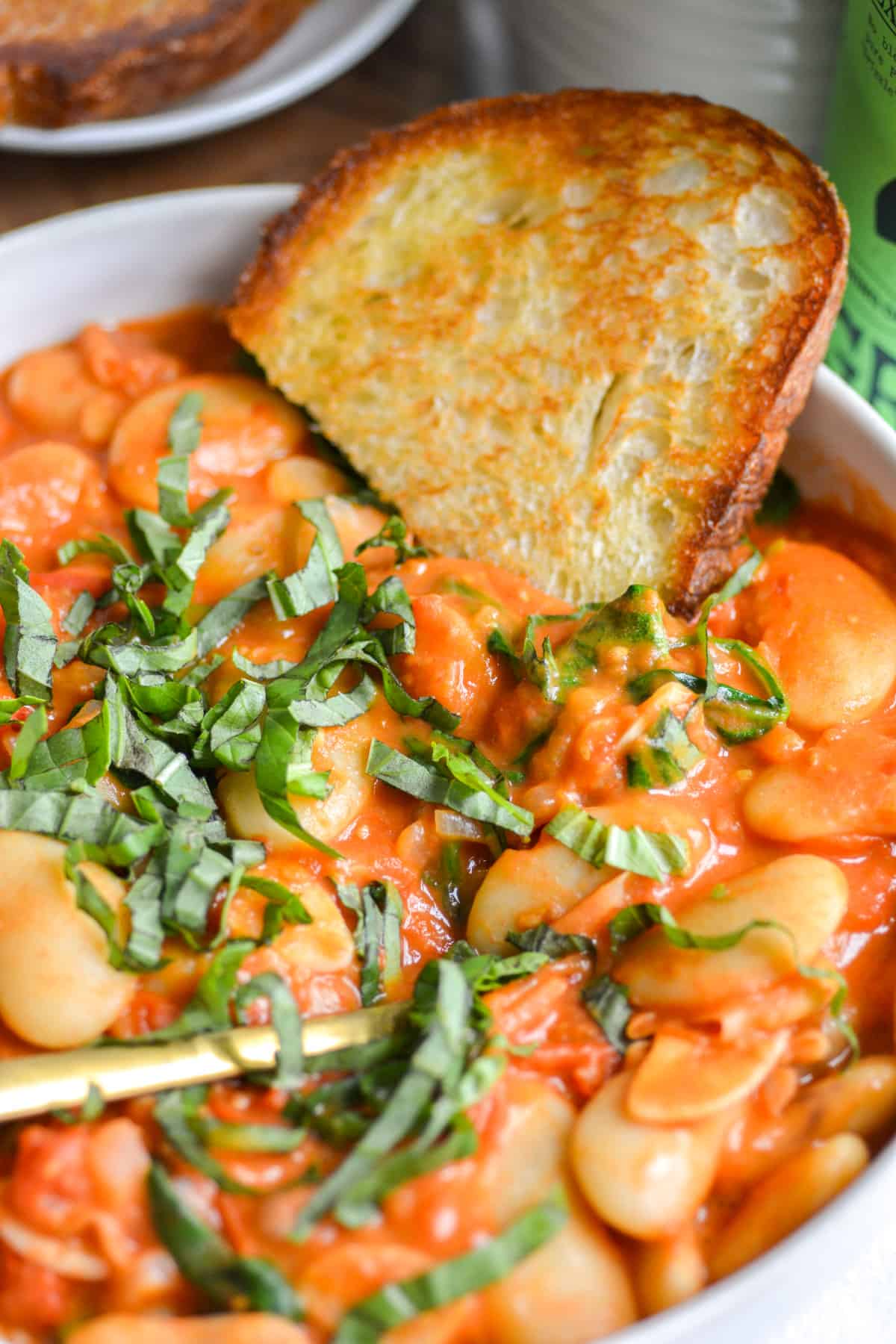 A piece of toast in a bowl of Creamy Vegetarian Tuscan Butterbeans that are topped with finely sliced basil.
