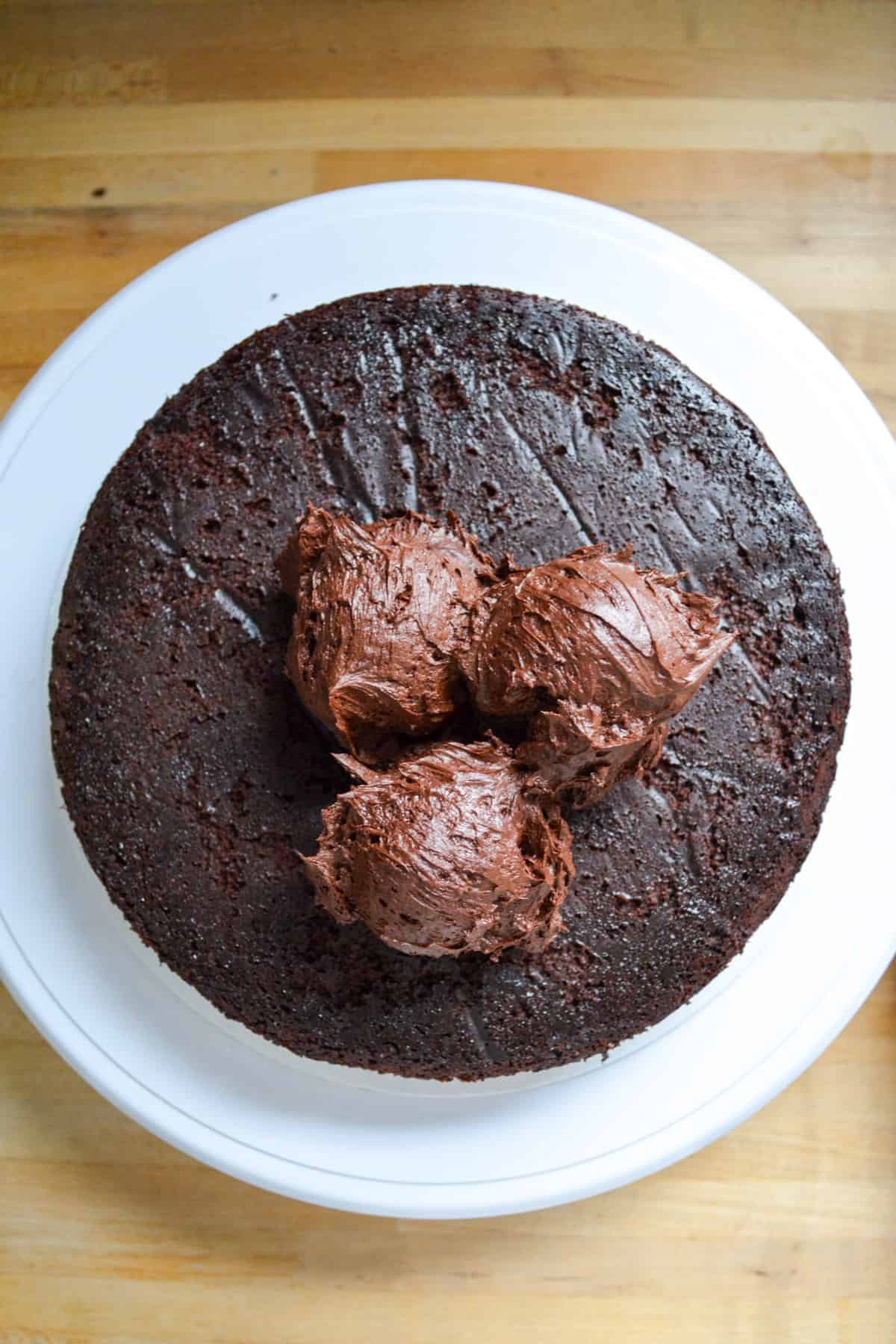 Vegan gluten free chocolate cake layer on a cake board with chocolate scoops of chocolate frosting on top of it.