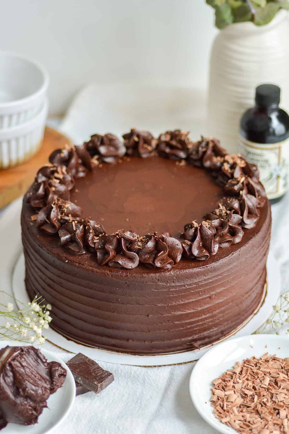 A round Vegan Gluten Free Chocolate Cake with shaved chocolate on top. Its placed on a cloth with a bowl of shaved chocolate in front.